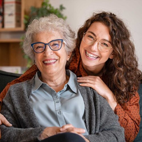 Portrait of old grandma and adult granddaughter hugging with love on sofa while looking at camera. Happy young woman with eyeglasses hugging from behind older grandma with spectacles. Senior woman spending time with her beautiful daughter, generation family concept.