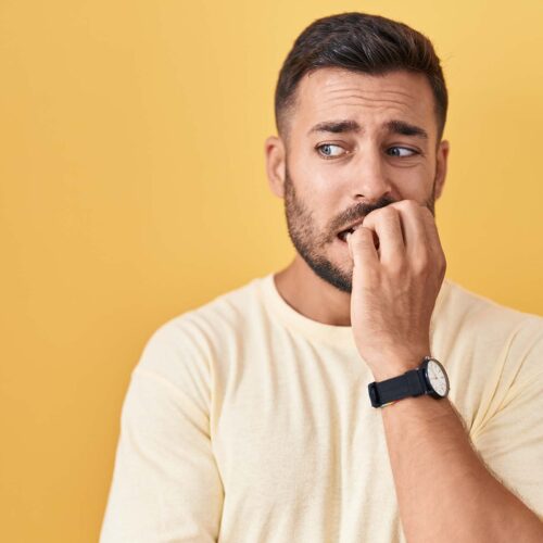 Handsome hispanic man standing over yellow background looking stressed and nervous with hands on mouth biting nails. anxiety problem.