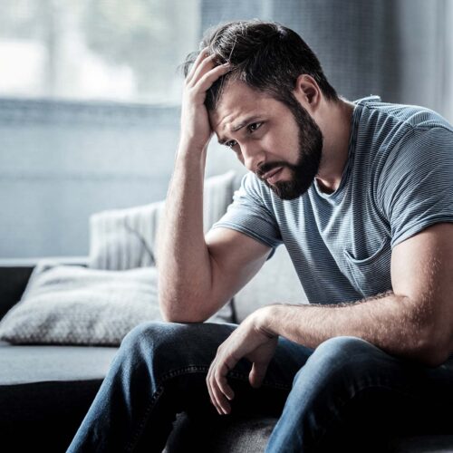 Unpleasant pain. Sad unhappy handsome man sitting on the sofa and holding his forehead while having headache