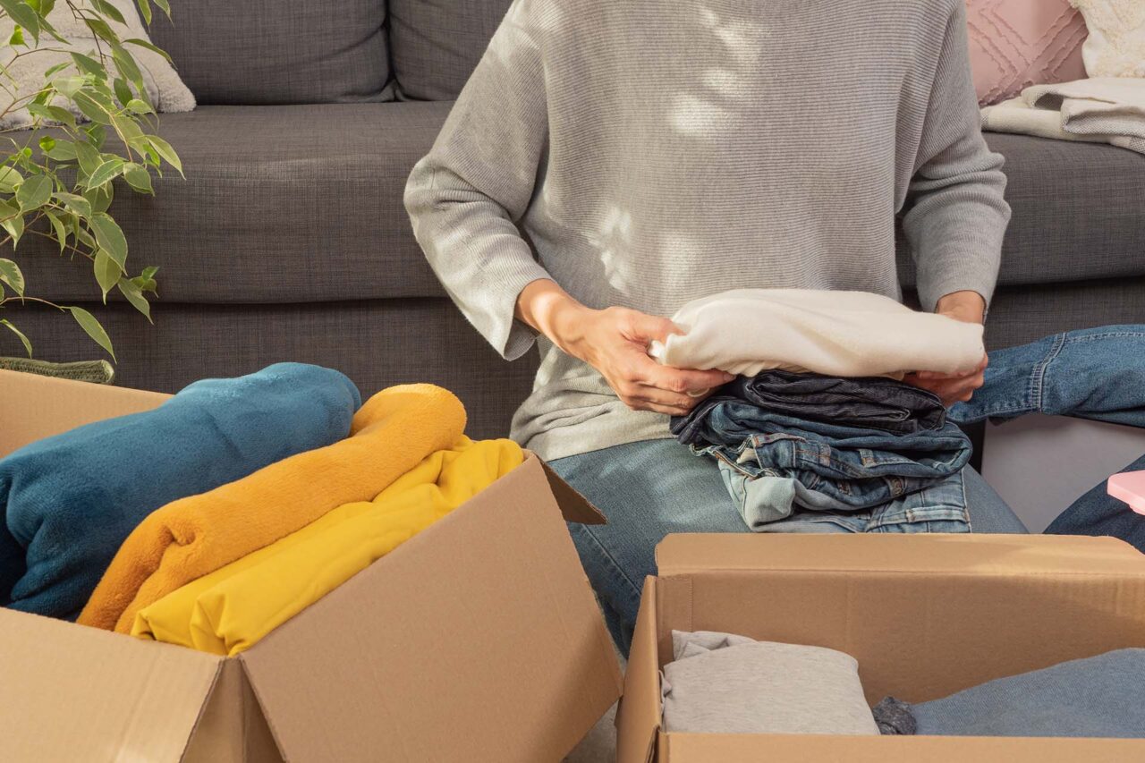 Woman sorting clothes and packing into cardboard box. Donations for charity, help low income families, declutter home, sell online, moving moving into new home, recycling, sustainable living concept.