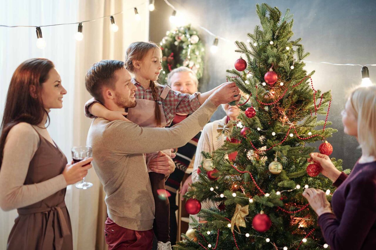 Young man with his little daughter decorating Christmas tree at home among other family members while preparing for holiday